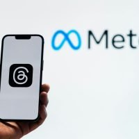 A hand holding a smartphone showing the Threads app with Meta logo in the background.